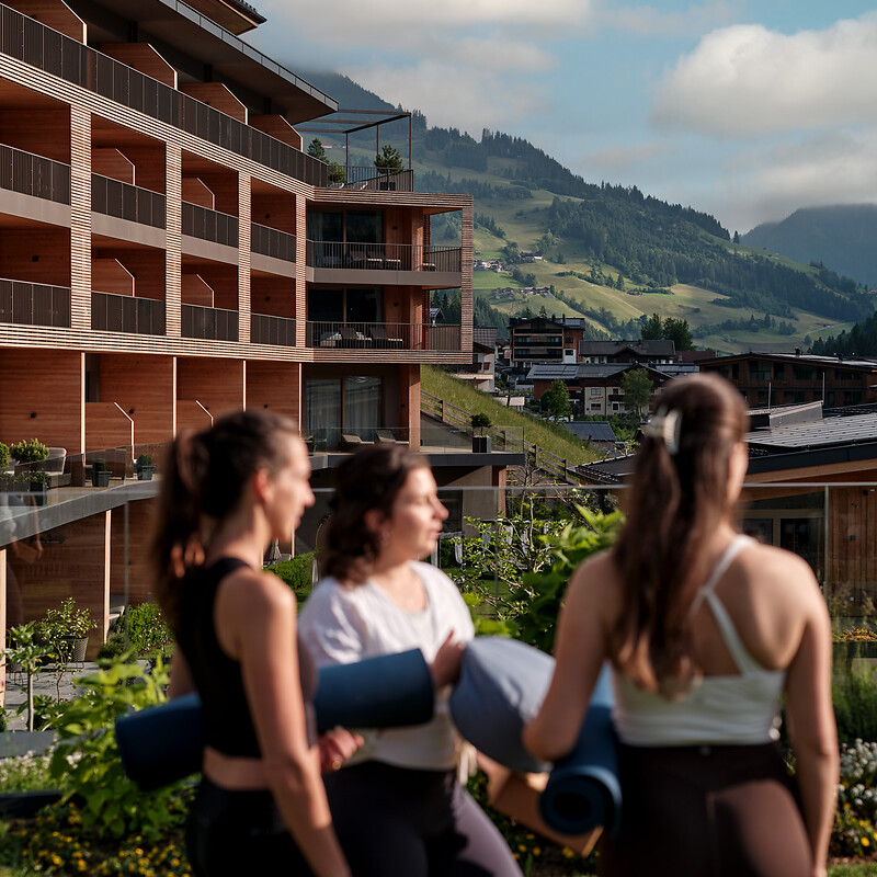Drei Frauen mit Yogamatten stehen vor einem modernen Holzgebäude in einer Berglandschaft.