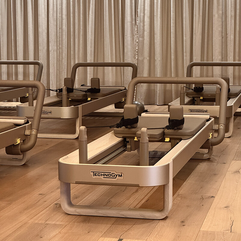 Mehrere Pilates-Reformer der Marke Technogym in einem Raum mit Holzboden und hellen Vorhängen.