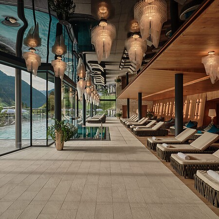 Langer Ruheraum mit Liegen, warmem Holzinterieur und dekorativen Hängelampen – links eine Glasfront mit Blick auf einen Pool und Berge.