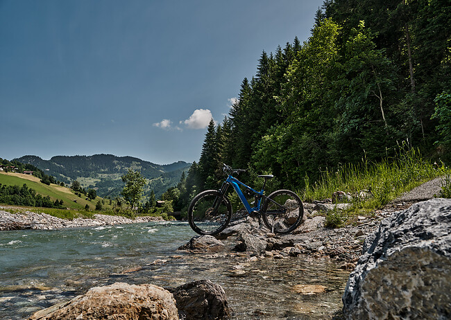 Mountainbike steht auf felsigem Ufer neben einem Fluss mit bewaldeten Hügeln im Hintergrund.