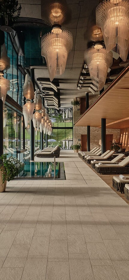 Langer Ruheraum mit Liegen, warmem Holzinterieur und dekorativen Hängelampen – links eine Glasfront mit Blick auf einen Pool und Berge.