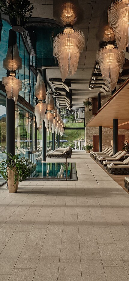 Langer Ruheraum mit Liegen, warmem Holzinterieur und dekorativen Hängelampen – links eine Glasfront mit Blick auf einen Pool und Berge.