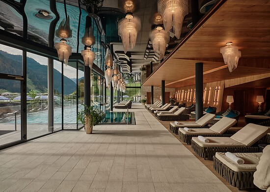 Langer Ruheraum mit Liegen, warmem Holzinterieur und dekorativen Hängelampen – links eine Glasfront mit Blick auf einen Pool und Berge.