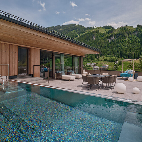 Moderner Holzbau mit großem Infinity-Pool und Terrasse mit Sitzgruppe vor bewaldetem Bergpanorama.