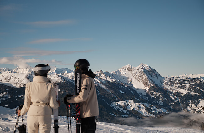 Zwei Skifahrer stehen nebeneinander im Schnee und blicken auf eine weite Berglandschaft mit schneebedeckten Gipfeln.