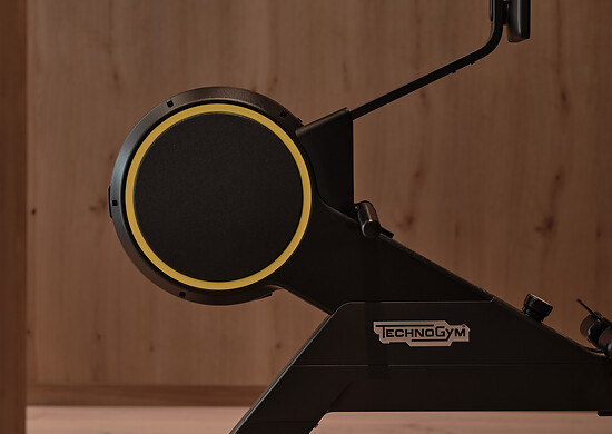 Seitliche Ansicht eines schwarzen Rudergeräts mit gelbem Ring und Technogym-Logo vor Holzwand.