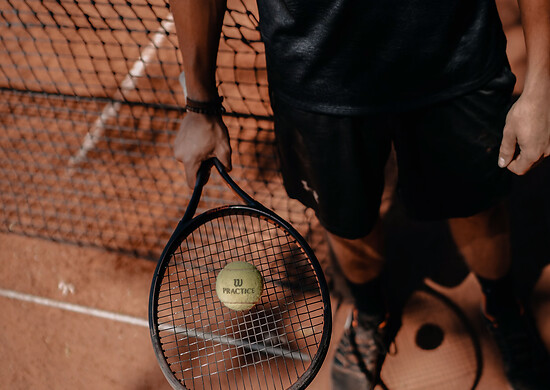 Tennisschläger mit Ball auf einem Tennisplatz, diesen hält eine Person in Sportkleidung.