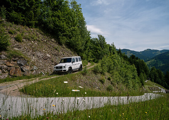 Geländewagen fährt auf schmalem Bergweg mit steilem Hang und grüner Vegetation unter bewölktem Himmel.