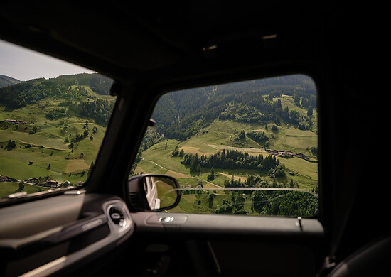 Aus dem Autofenster heraus ist eine grüne Berglandschaft mit Wiesen, vereinzelten Häusern und bewaldeten Hängen zu sehen - der Innenraum des Fahrzeugs ist ebenfalls noch teilweise zu sehen.