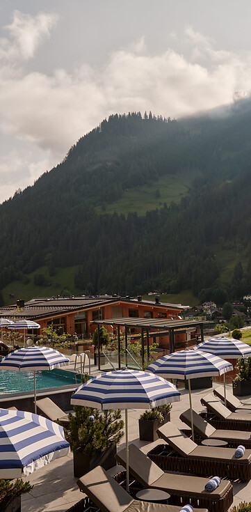 Hotelterrasse mit Liegestühlen und blau-weiß gestreiften Sonnenschirmen vor Bergkulisse.