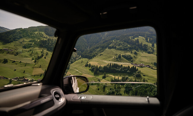 Aus dem Autofenster heraus ist eine grüne Berglandschaft mit Wiesen, vereinzelten Häusern und bewaldeten Hängen zu sehen - der Innenraum des Fahrzeugs ist ebenfalls noch teilweise zu sehen.
