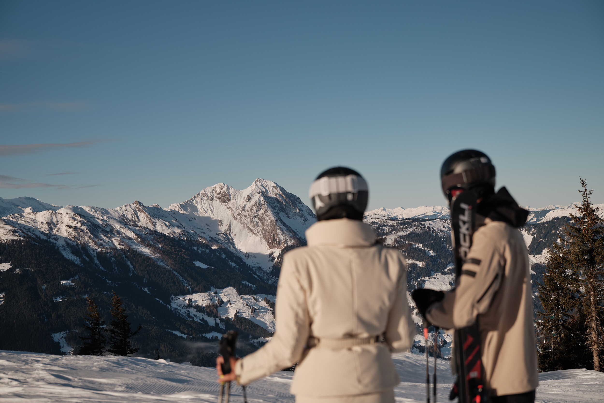 Ski In Ski Out: couple with skis in hand in beige, black ski suit with panoramic views of the Großglockner in Salzburger Land